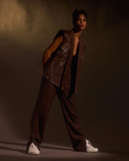 A woman in dark brown pants, a sleeveless leather vest, and white sneakers stands against a neutral background, with her hands in her pockets and a serious expression.