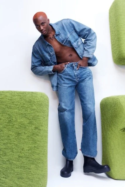 Person with short hair wearing a denim jacket open to reveal bare chest and wearing jeans, standing with one hand in pocket amidst green padded furniture pieces.