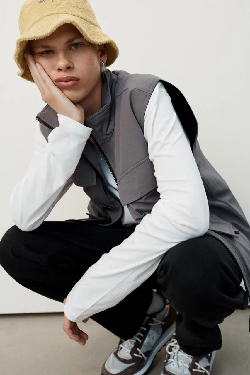 A young man wearing a beige bucket hat, gray vest over a white long-sleeve shirt, black pants, and patterned sneakers, squatting and resting his face on his hand while looking at the camera.