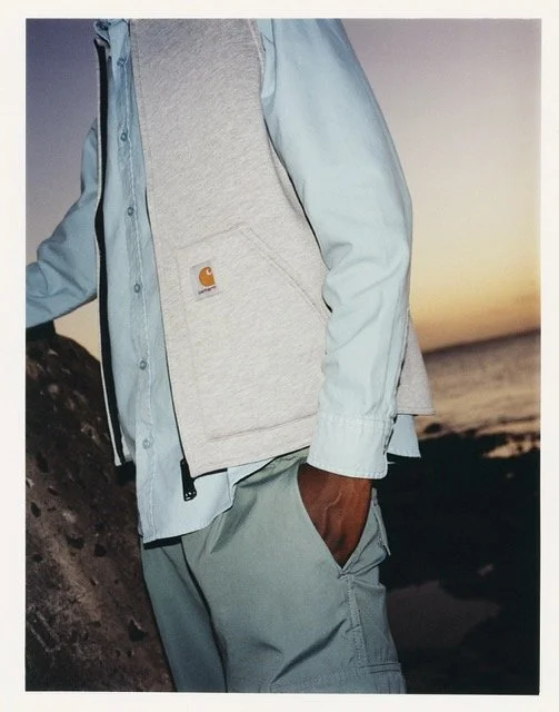Person wearing a light blue jacket and a gray Carhartt hoodie, standing outdoors at sunset near a rocky surface.