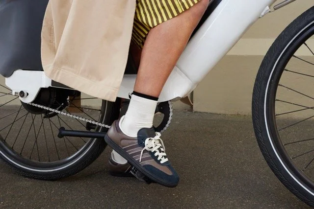 Person in a wheelchair wearing gray sneakers, beige pants, yellow striped shirt, and white socks, with a beige coat hanging from the wheelchair.
