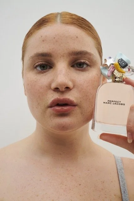 A woman with red hair and freckles holding a perfume bottle labeled 'Perfect Marc Jacobs,' with a playful bow-shaped decoration on the cap.