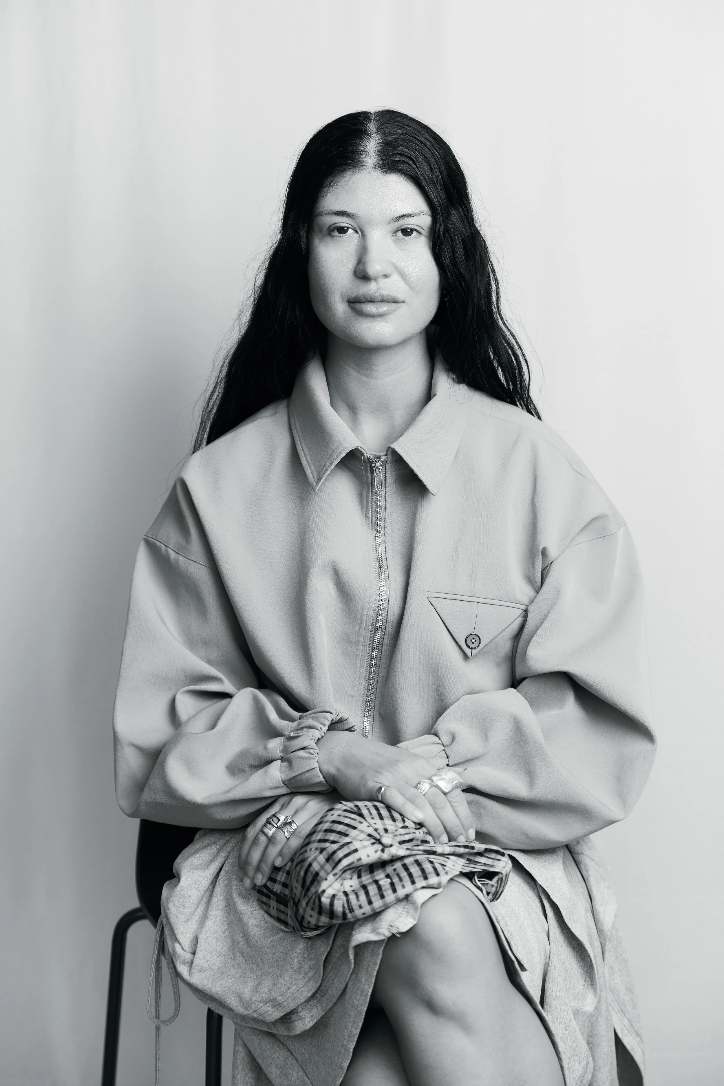 Black and white portrait of Danielle Soglimbene with long dark wavy hair, sitting on a stool against a plain light-colored background. She is wearing a zip-up jacket and a plaid garment on her lap.