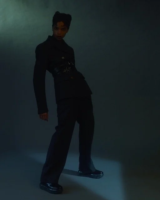 A woman in a black suit and platform shoes standing in front of a plain dark background, looking at the camera.