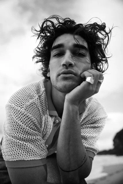 Black and white photo of a young man with messy hair, resting his chin on his hand, outdoors with cloudy sky behind him.