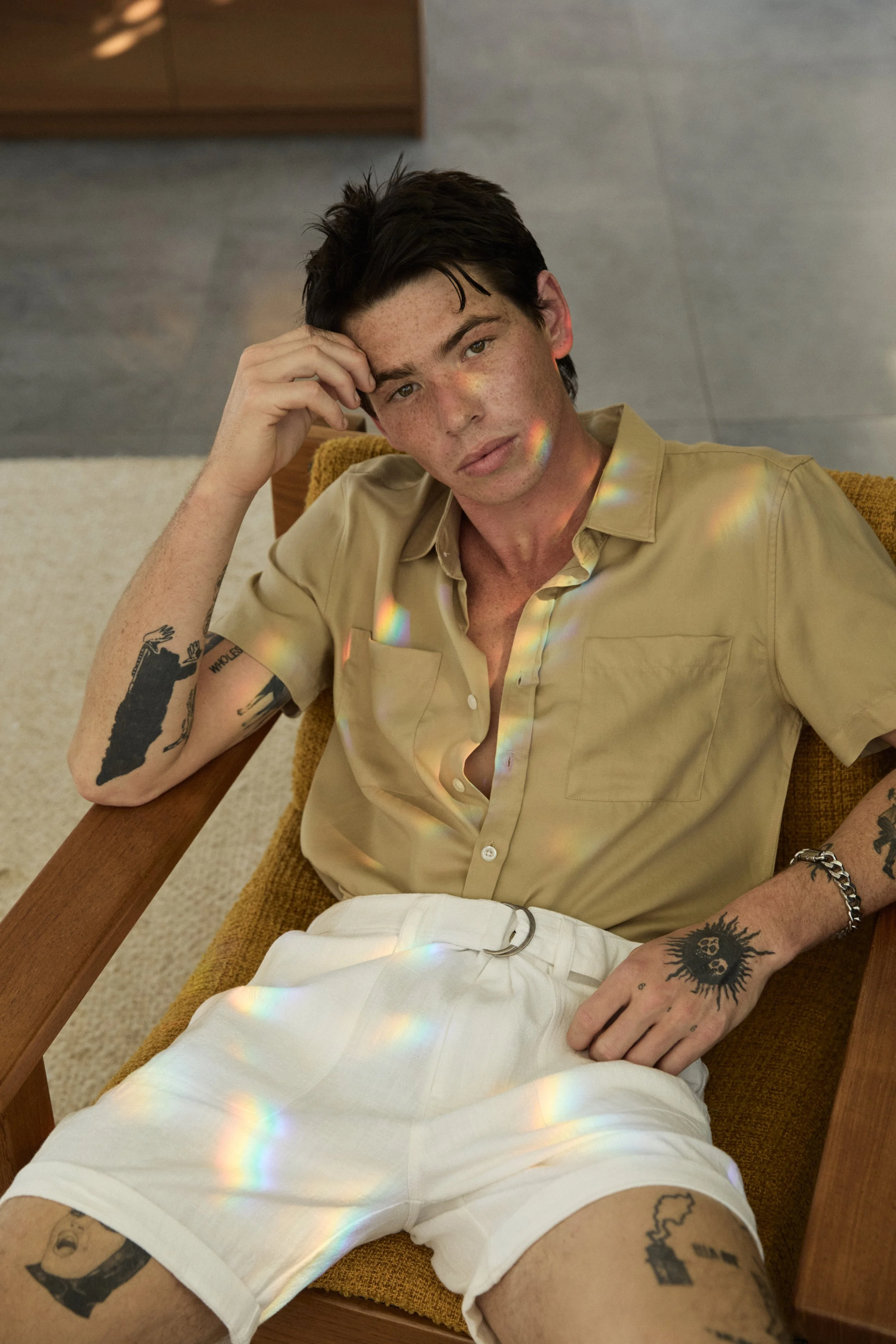 A young man with dark hair, freckles, and tattoos on his arms and legs, sitting in a brown chair with a yellow cushion. He's wearing a beige short-sleeved shirt and white shorts. There are rainbow reflections on his face and clothing, and he is resti