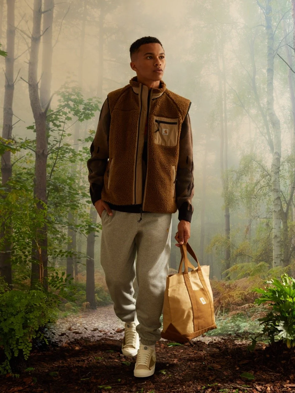 A person standing on a forest trail holding a tote bag, wearing a brown vest, gray sweatpants, and white sneakers, with trees and greenery in the background.