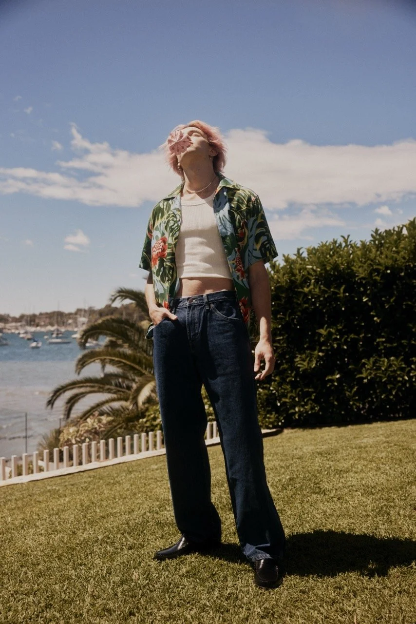 A woman with pink hair stands outdoors on a grassy area near water and boats, wearing a floral shirt over a white crop top, dark jeans, and black shoes, with her face partially obscured by a pink flower.