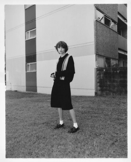 A young person with curly hair standing on a grassy lawn in front of an apartment building, wearing a black dress, jacket, and loafers with socks, holding a small object in their hand.