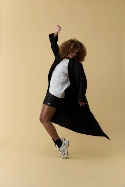 Person with curly hair dancing against a beige background, wearing a black coat, black shorts, a white shirt, and athletic shoes.