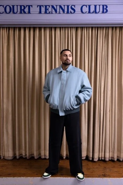 Man standing in front of beige curtain with 'Court Tennis Club' sign above, wearing a light blue jacket, black pants, and sneakers.