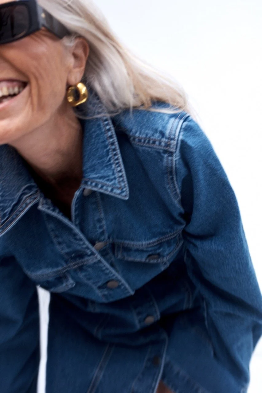 Close-up of a woman smiling, wearing a denim jacket, gold earrings, and sunglasses with her hair partially visible.