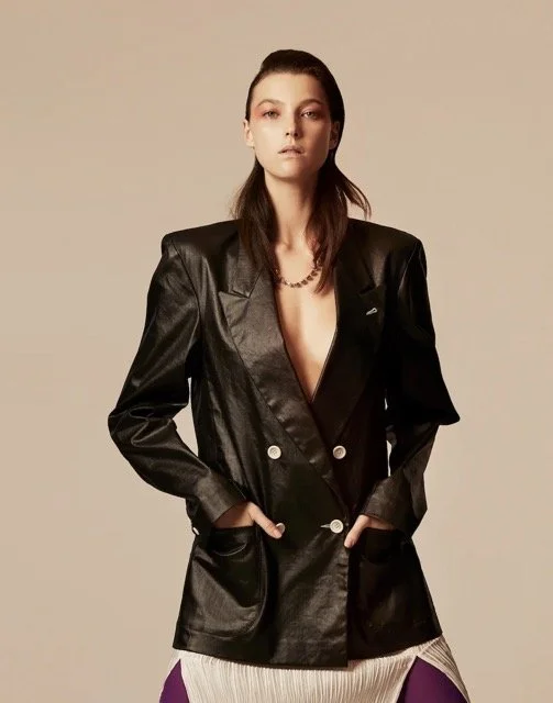 Fashion model wearing a black leather blazer over a low-cut top, accessorized with a necklace, posing with hands in blazer pockets against a beige background.