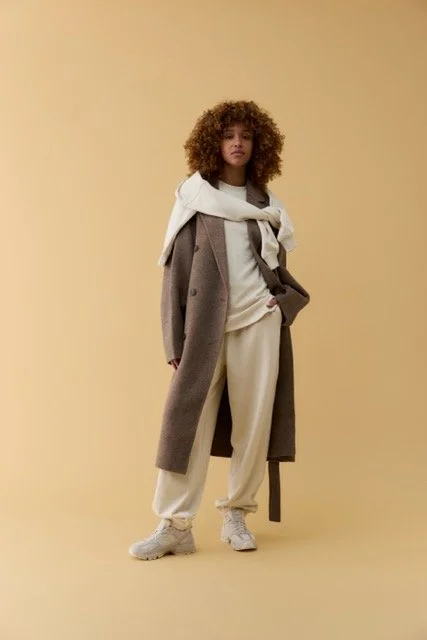 Woman with curly hair wearing a beige hoodie, gray coat, beige sweatpants, and sneakers standing against a plain beige background.