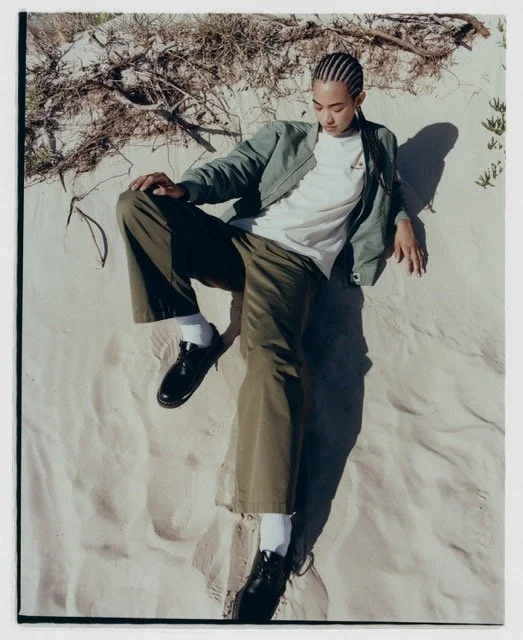 Person lying on sand at the beach, wearing a green jacket, white shirt, brown pants, black shoes, and white socks, with braided hair and arms outstretched.