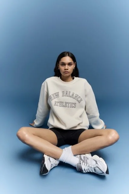 Young woman sitting on the floor against a blue background, wearing a white sweatshirt, black shorts, white socks, and sneakers.