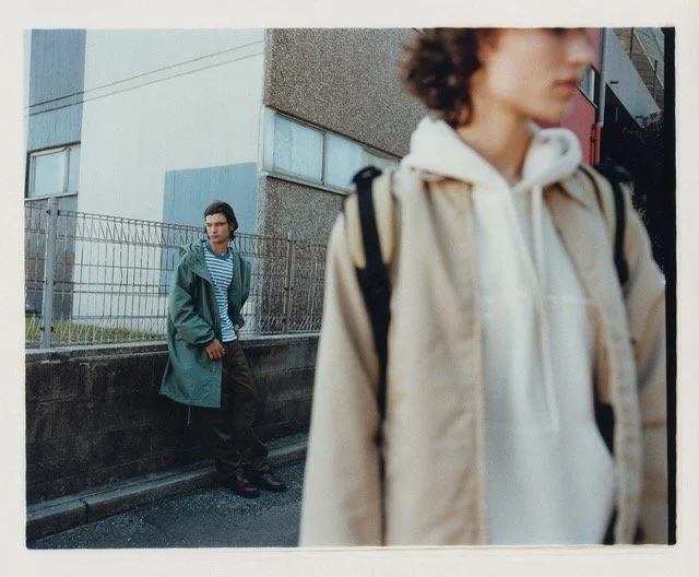 Two young people standing outdoors in an urban area; one in the foreground facing away, wearing a beige jacket and hoodie, with a backpack, and the other in the background leaning against a wall, wearing a green jacket and striped shirt.