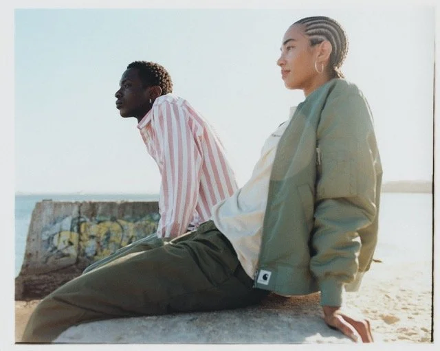 Two young adults sitting on a concrete ledge outdoors near the water, with a graffiti-covered wall behind them. The woman is wearing a green jacket and the man is in a striped shirt.