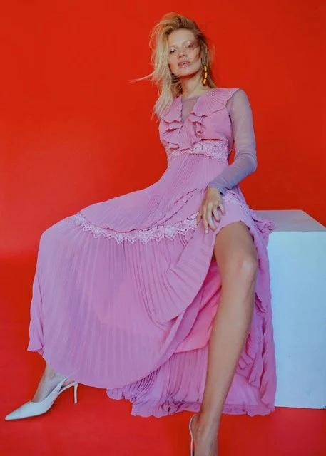 Woman in a pink pleated dress with ruffles, sitting on a white block against a red background, wearing white high heels and gold earrings.