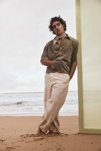 Young man with curly hair leaning against a wall at the beach, wearing a brown shirt and light-colored pants.