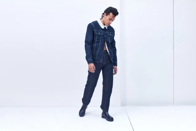 A man dressed in a denim jacket, dark pants, and black shoes standing against a plain white background.