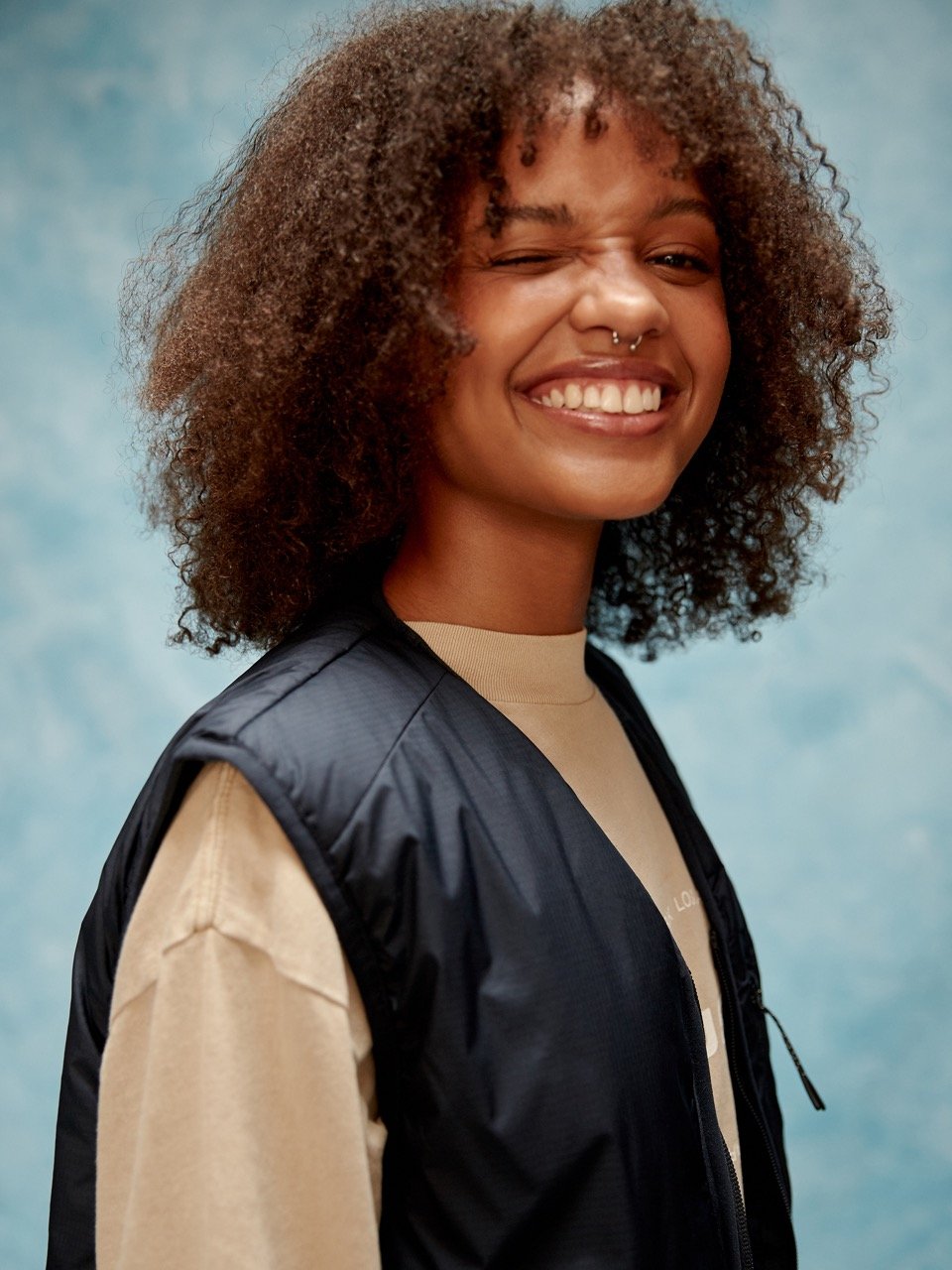 Smiling young woman with curly hair, wearing a black vest over a beige shirt, winking at the camera.