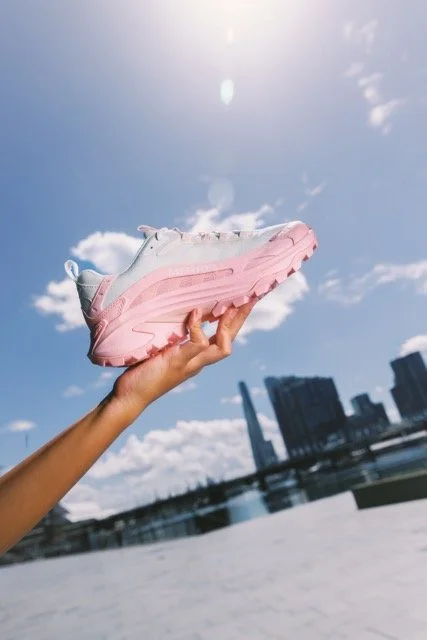 Hand holding a white and pink sneaker with a clear blue sky and city skyline in the background.