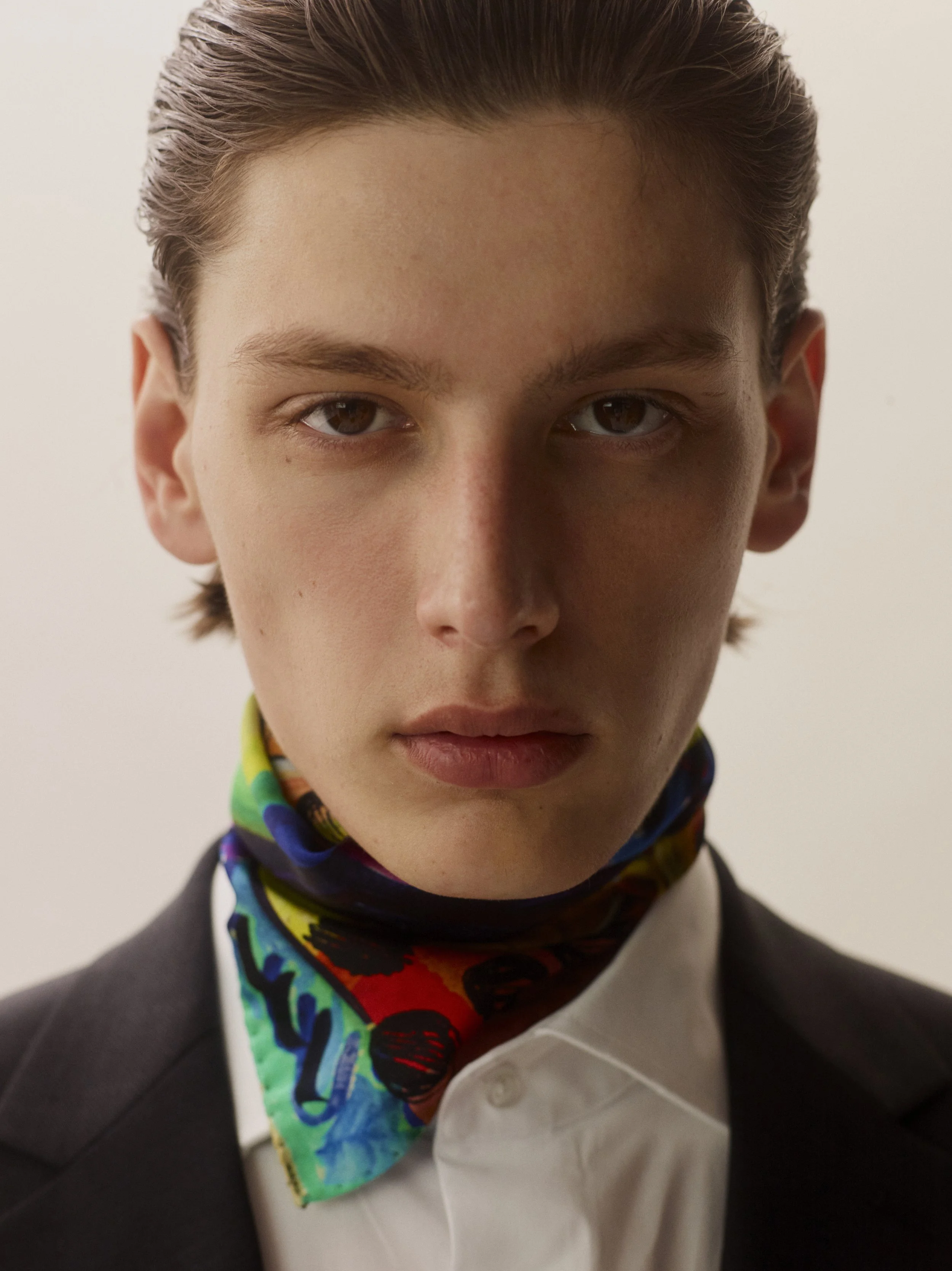 Close-up of a young man with slicked-back hair, wearing a black suit, white shirt, and a colorful, patterned scarf around his neck.