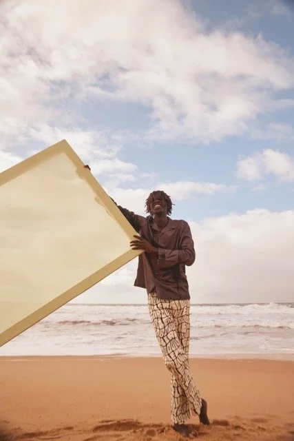 Person standing on a beach holding a large piece of surfboard, with ocean waves and a cloudy sky in the background.