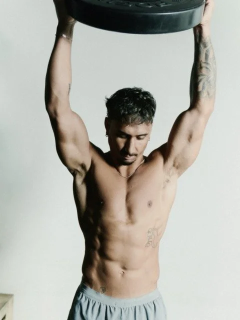 A shirtless man with tattoos lifting a large black weight plate overhead.