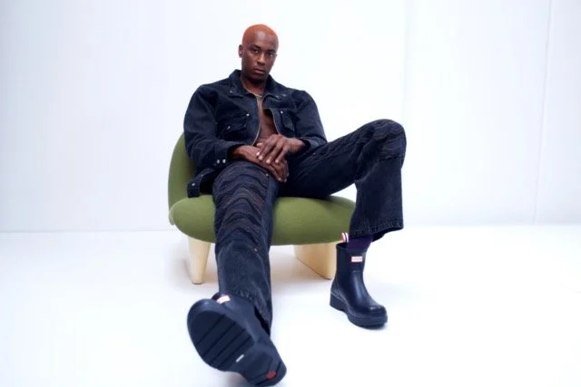 A man sitting in a green chair with a plain white background, wearing a black jacket, dark jeans, and black boots, with one leg extended toward the camera.