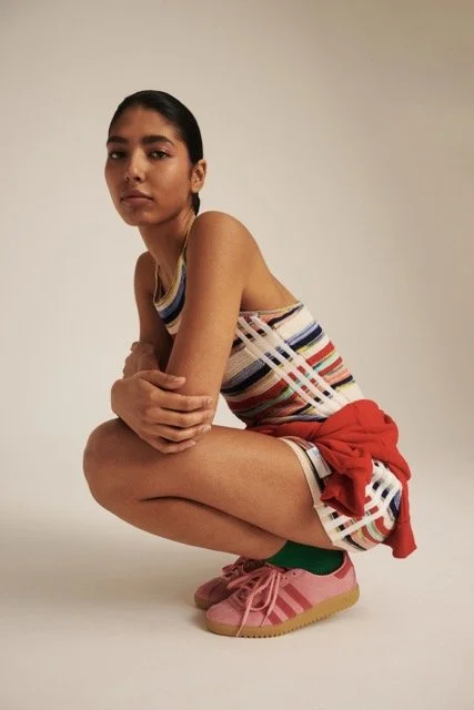Young woman crouching in studio, wearing striped sleeveless top, red jacket tied around waist, pink sneakers, and green socks.