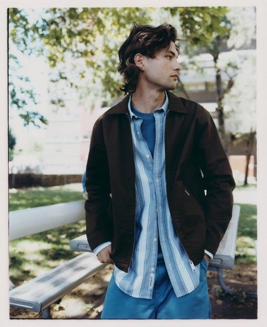 Young man standing outdoors by park benches, wearing a black jacket over a blue striped shirt and blue pants, with curly dark hair.
