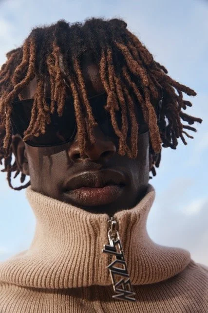 Young man with dreadlocks, sunglasses, and a zip-up beige sweater with a large 'V' zipper pull, outdoors against a blue sky.