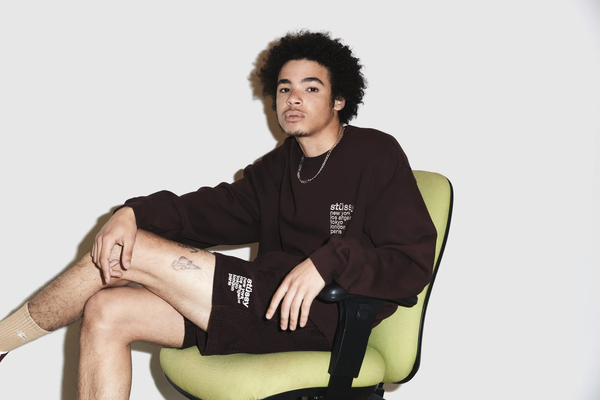 Young man sitting on a green office chair, wearing a black sweatshirt with city names, shorts, and socks, with tattoos on his legs, against a plain background.