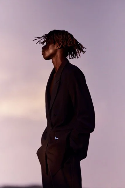 A man with dreadlocks wearing an oversized black blazer against a pastel-colored sky.