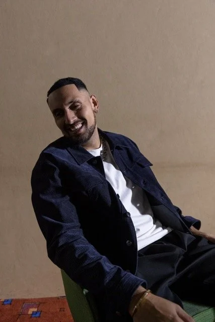 Man sitting on a green chair, smiling, wearing a dark jacket over a white shirt, against a plain beige wall.