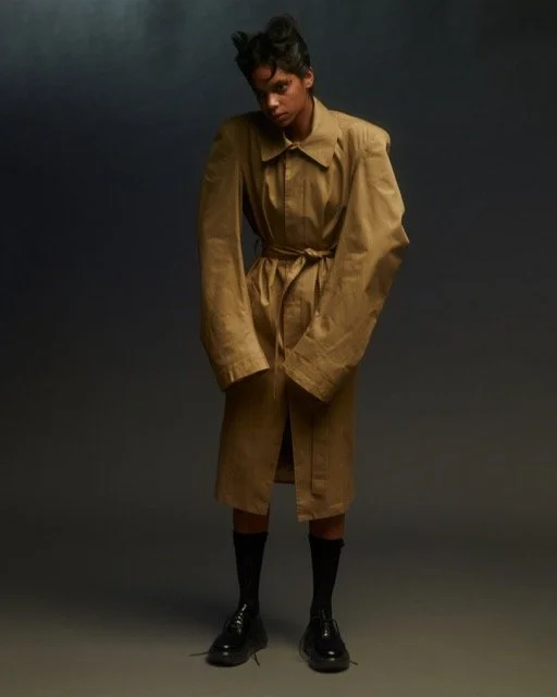 Young woman in a long tan trench coat, black knee-high socks, and black shoes standing against a dark gray background.