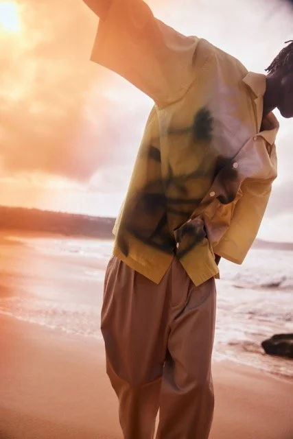 Person standing on the beach at sunset, wearing a beige jacket and brown pants.