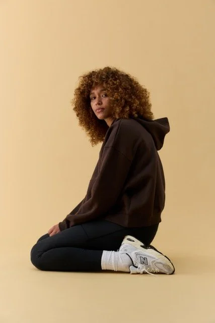 A young woman with curly hair in a black hoodie and black pants kneeling on the floor against a beige background.