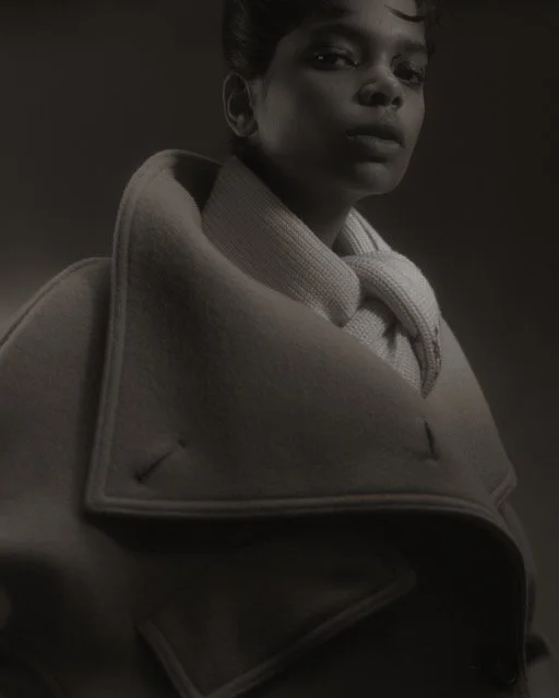 A black and white portrait of a woman looking at the camera, wearing a coat with the collar turned up.