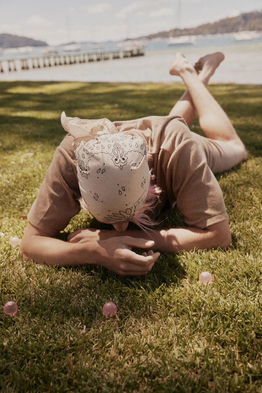 Person lying on grass near a body of water with boats and a shoreline in the background, wearing a bandana on their head.
