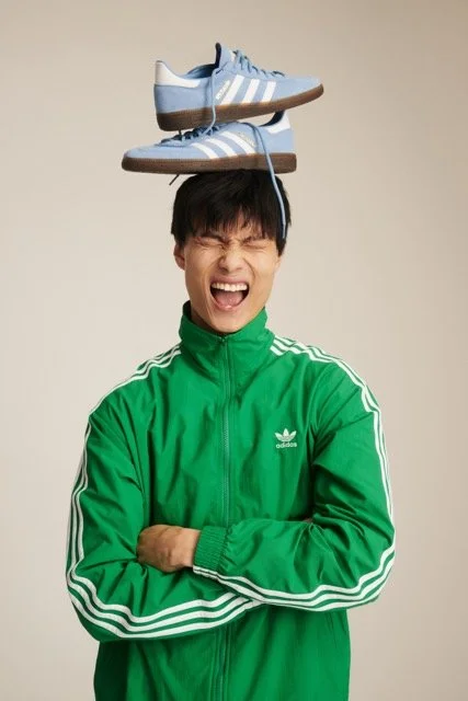 Young man in a green Adidas track jacket with arms crossed, balancing a pair of light blue and white sneakers on his head, eyes closed, and mouth open in a playful expression.