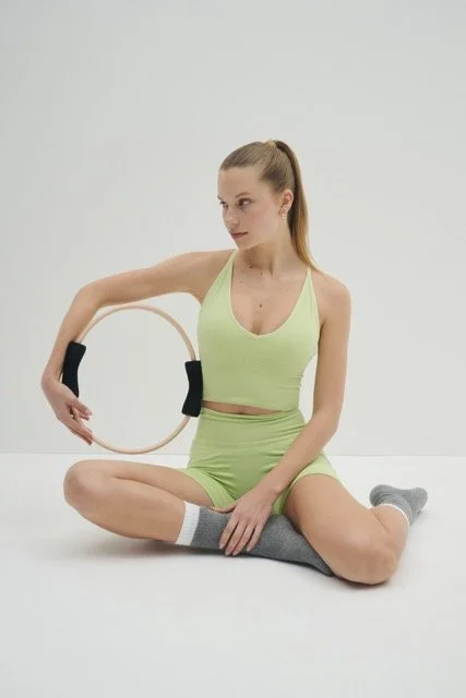 Young woman in light green athletic wear sitting cross-legged on the floor holding a Pilates ring.