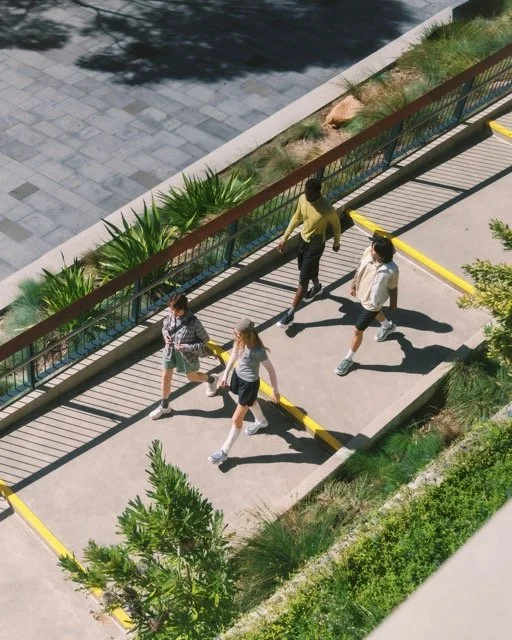 Four children walk on a sidewalk near a parking lot, holding hands and wearing school uniforms, with a man walking beside them.