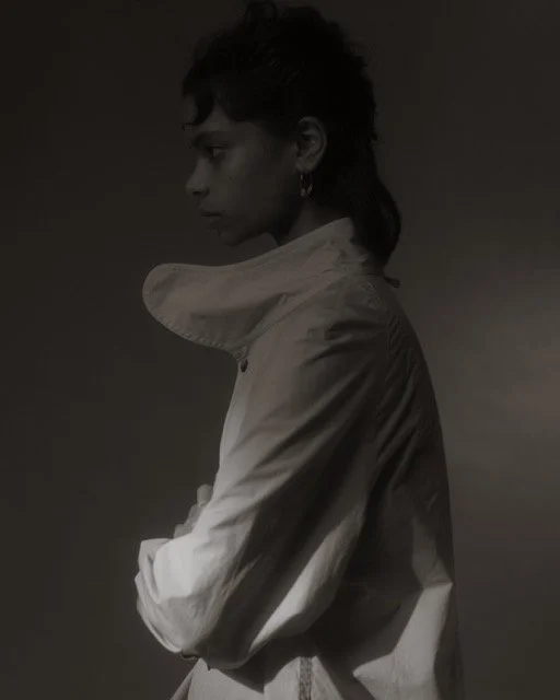 Black and white photo of a person with short, curly hair, wearing a high-collared jacket, looking to the side with crossed arms.