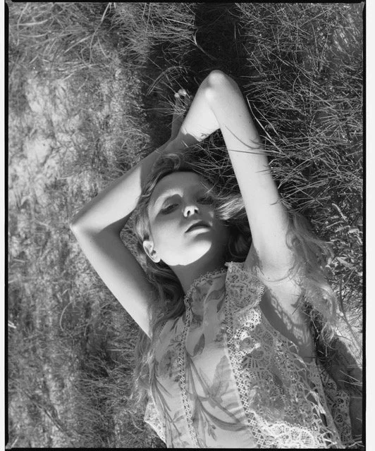 A woman lying on the grass with her arms raised above her head, wearing a lace dress, in black and white.