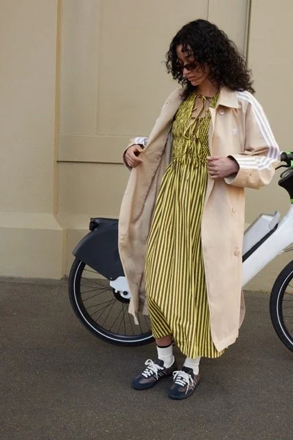 A woman with curly hair and glasses standing next to an electric bicycle, wearing a yellow and black striped dress, a beige Adidas jacket, and black sneakers.