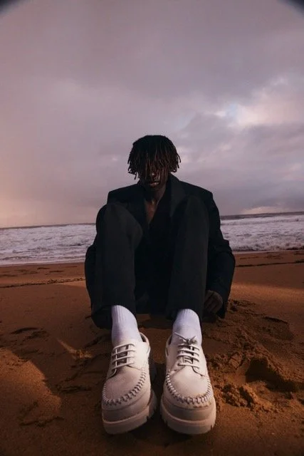 Person sitting on a sandy beach at sunset with large white boots front and center, dark jacket, and dreadlocks, with ocean waves and cloudy sky in the background.