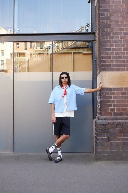 Person wearing sunglasses, a blue shirt over a white T-shirt, black shorts, striped socks, and sneakers, standing against a brick wall with a glass window behind them.